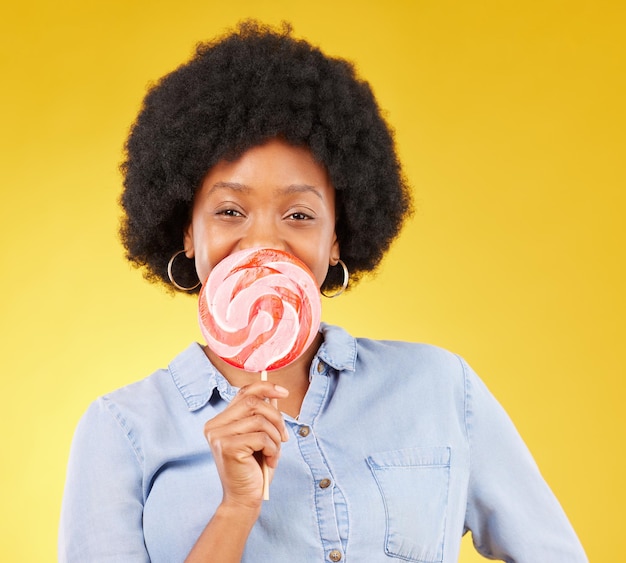 Dolci caramelle e lecca-lecca con donna nera in studio per colorato allegro e positivo Giovane felicità e dessert con femmina isolata su sfondo giallo per dolcetti alimentari e dolciumi