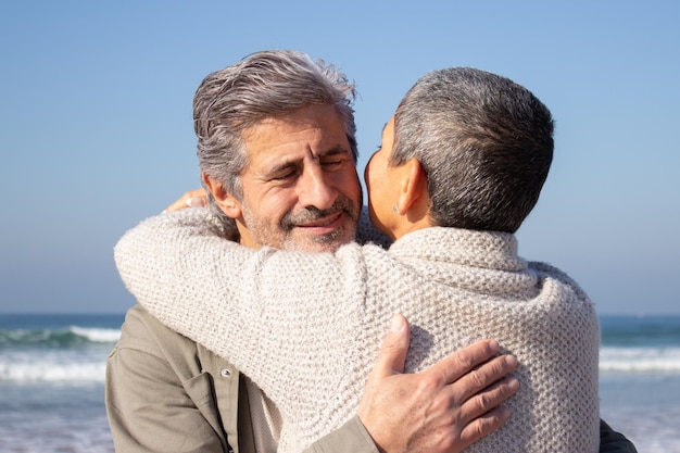 Dolci coppie senior che si abbracciano in riva al mare in una soleggiata giornata autunnale. Signora dai capelli corti e uomo anziano barbuto che si abbracciano con cura. Relazione, amore, concetto di viaggio