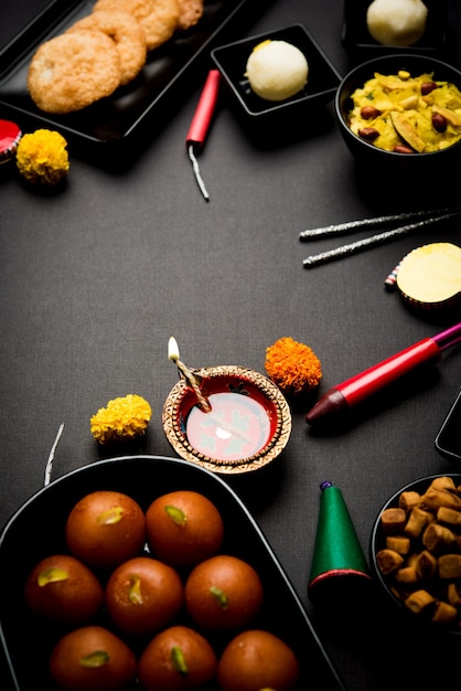 Dolci e snack Diwali disposti in un gruppo con Diya o lampada a olio, fiori e petardi o Patakhe su una superficie lunatica, messa a fuoco selettiva