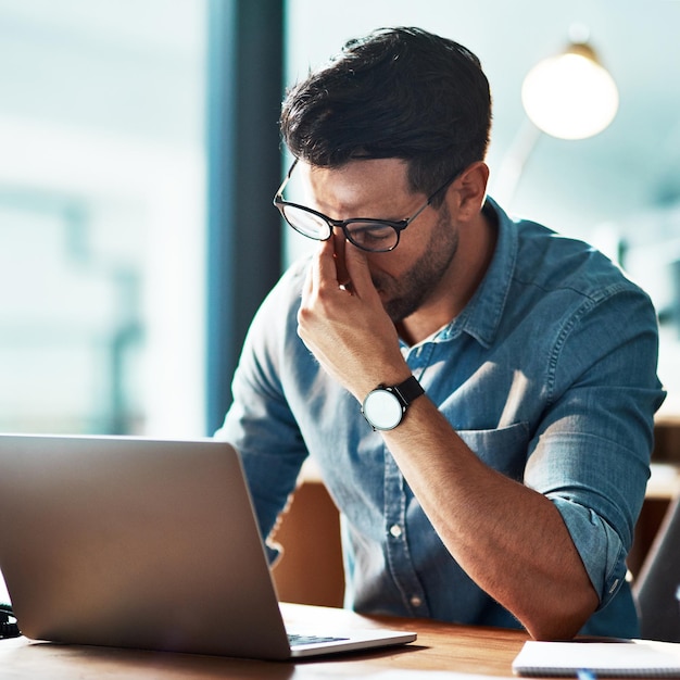 Dolore da stress e affaticamento degli occhi dovuto alle lunghe ore di lavoro sul laptop che causano mal di testa problemi di vista e vista stanchi Burnout giovane uomo d'affari stanco e sovraccarico di lavoro che rimuove gli occhiali per strofinare gli occhi asciutti