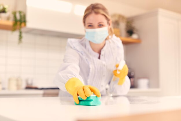 Donna a casa che indossa una maschera in cucina che fa i lavori domestici e pulisce la superficie del bancone con spray