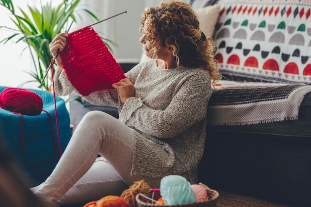 Donna a casa seduta sul tappeto facendo lavori a maglia con lana colorata. Autunno e inverno vestiti caldi realizzati a mano da persone di sesso femminile che si godono l'hobby e il tempo libero al coperto