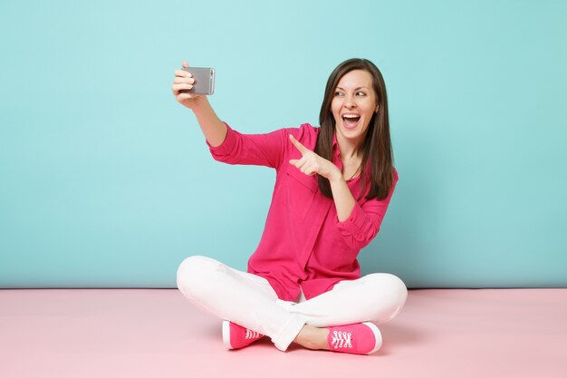 Donna a figura intera in camicia rosa, pantaloni bianchi seduta sul pavimento facendo selfie sul cellulare