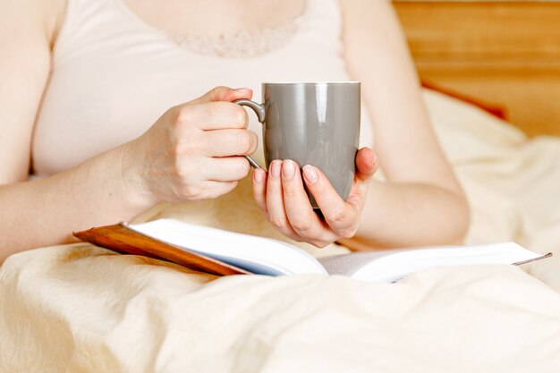 Donna a letto con una tazza di tè e un libro. Lettura della donna adulta nel letto.