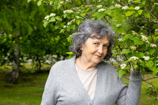 Donna abbastanza anziana in età pensionabile che cammina nel parco tra alberi in fiore. Ritratto di una bella donna dai capelli grigi in un parco con alberi in fiore. Primavera e tanti alberi verdi