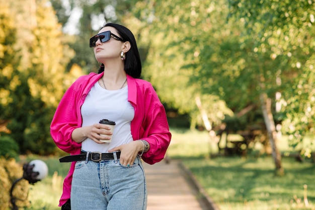 Donna abbastanza castana in occhiali da sole con una tazza di caffè che cammina nel parco