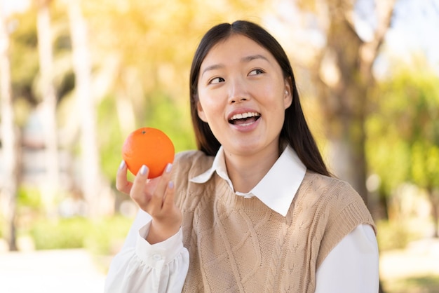 Donna abbastanza cinese all'aperto che tiene un'arancia con l'espressione felice