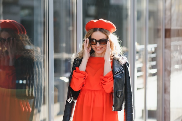 Donna abbastanza felice con un dolce sorriso in occhiali da sole vintage in cappello in abiti casual rosso-neri con caffè passeggiate e sorrisi all'aperto in città