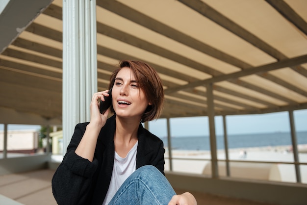 Donna abbastanza giovane allegra che parla sul telefono cellulare sulla terrazza sulla spiaggia