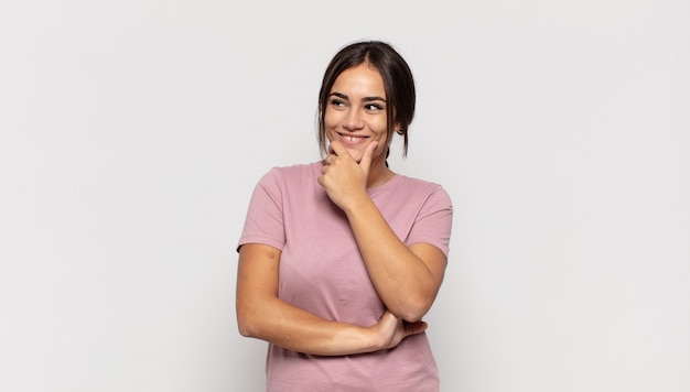 Donna abbastanza giovane che sorride con un'espressione felice e sicura con la mano sul mento, chiedendosi e guardando di lato