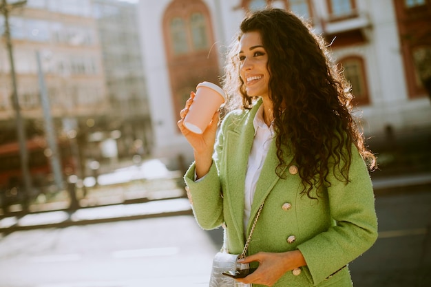 Donna abbastanza giovane che utilizza smartphone per strada e tiene caffè da asporto