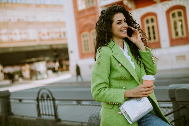 Donna abbastanza giovane che utilizza smartphone per strada e tiene caffè da asporto