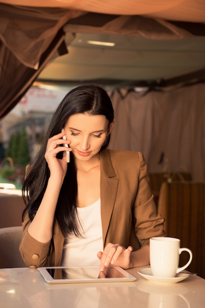 Donna abbastanza giovane di affari in un caffè parlando al telefono con tablet