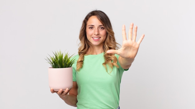 Donna abbastanza magra che sorride e sembra amichevole, mostra il numero cinque e tiene in mano una pianta decorativa