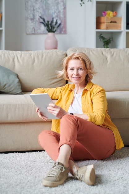 Donna abbastanza matura con tavoletta digitale rilassante sul pavimento dal divano nel tempo libero o durante il periodo di auto isolamento domestico