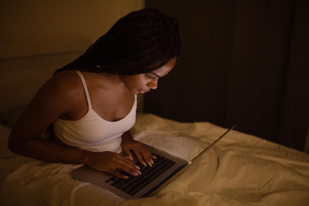 Donna abbastanza nera felice che utilizza il computer portatile sul letto. Donna che utilizza laptop di notte.