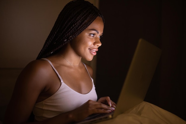 Donna abbastanza nera felice che utilizza il computer portatile sul letto. Donna che utilizza laptop di notte.