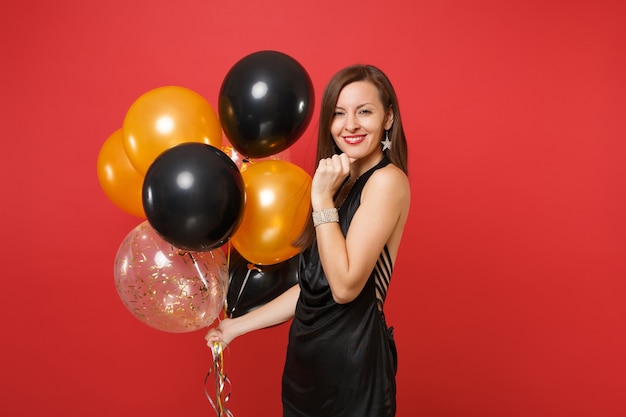 Donna abbastanza sorridente in abitino nero che celebra tenendo la mano sul mento tenendo i palloni ad aria isolati su sfondo rosso. San Valentino, felice anno nuovo, concetto di festa di compleanno mockup.