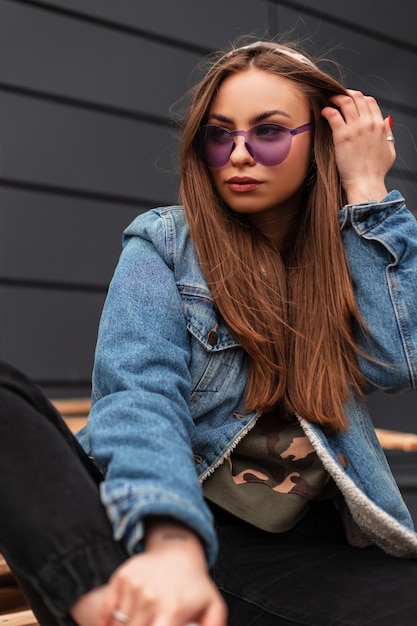 Donna abbastanza urbana giovane hipster raddrizza i capelli che si siede su tavole di legno. Ragazza attraente in vestiti di jeans alla moda della gioventù in occhiali viola moda in posa su pallet vintage vicino al muro grigio della città