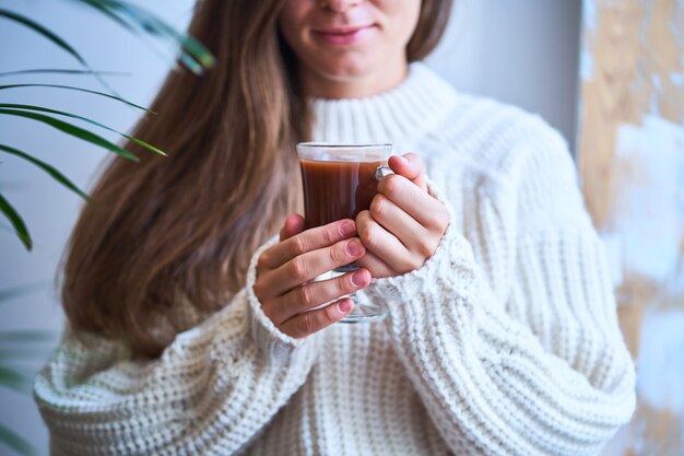 Donna accogliente in maglione bianco lavorato a maglia che tiene un bicchiere di cacao dolce riscaldante. Bevande calde