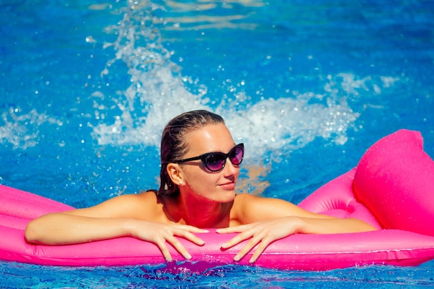 Donna adatta che lavora in piscina. Giovane bella ragazza che fa esercizio di acquagym galleggiante su un materasso rosa gonfiabile spruzza acqua spray