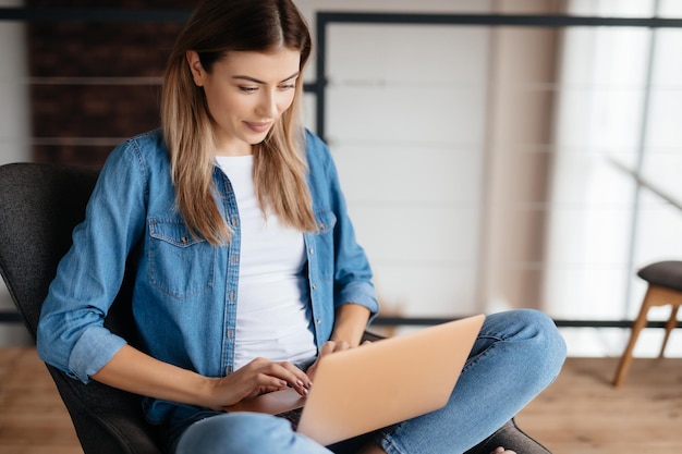 Donna adorabile concentrata che sorride e guarda lo schermo del laptop mentre è seduto a gambe incrociate in poltrona