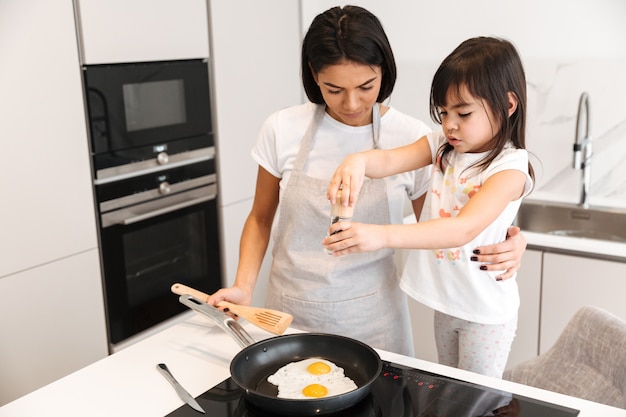 Donna adorabile del brunette in grembiule che cucina e che frigge insieme le uova con la sua piccola figlia in cucina