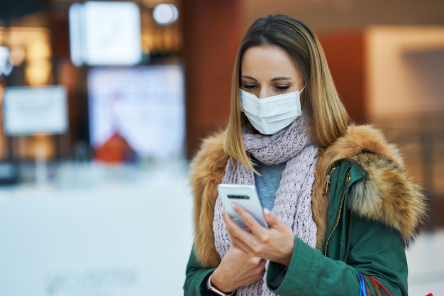 donna adulta che fa shopping nel centro commerciale utilizzando lo smartphone che indossa una maschera, concetto di coronavirus