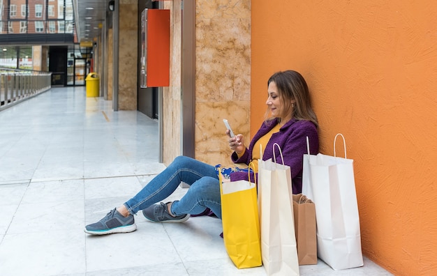 Donna adulta che si siede sul pavimento guardando il telefono cellulare con le borse della spesa accanto a lei