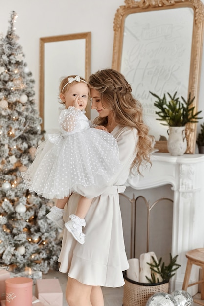 Donna adulta con un tenero trucco e capelli lunghi in posa con la figlia carina del bambino in interni natalizi decorati.