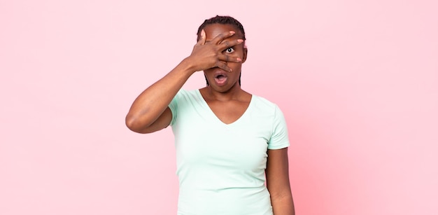 Donna adulta nera afro che sembra scioccata, spaventata o terrorizzata, coprendo il viso con la mano e sbirciando tra le dita