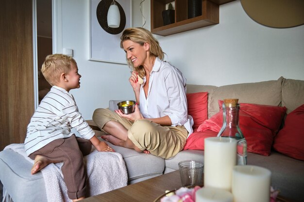 Donna adulta sorridente con figlio piccolo seduto sul divano nel soggiorno di casa e facendo colazione