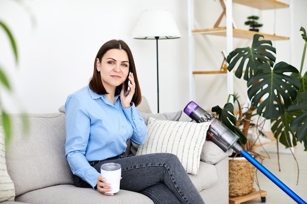 Donna affascinante che parla sullo smartphone seduto sull'accogliente divano di casa e tiene una tazza in mano La giovane donna sorridente che chiacchiera con un amico sul telefono cellulare ha piacere di una conversazione telefonica