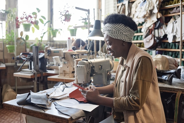 Donna africana che legge un messaggio sul cellulare mentre cuce vestiti in fabbrica