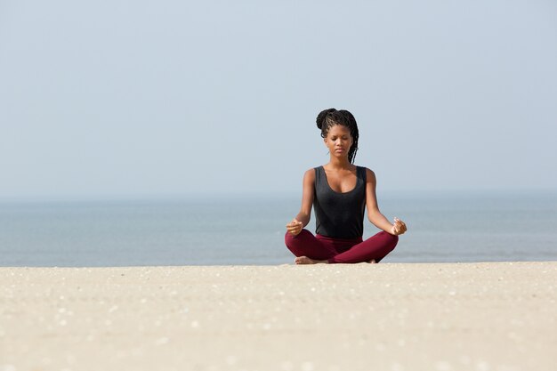 Donna africana che si siede alla spiaggia nella posa di yoga