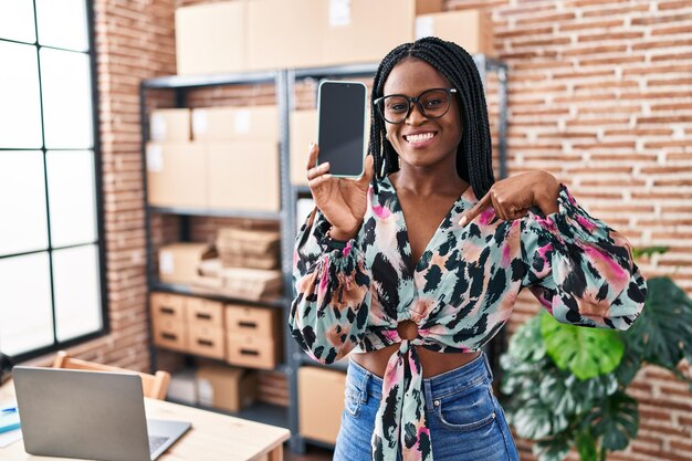 Donna africana con trecce che lavora presso un e-commerce di piccole imprese che mostra lo schermo dello smartphone che punta il dito verso un auto sorridente felice e orgoglioso