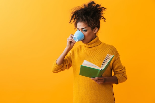 Donna africana confusa che indossa un maglione in piedi isolato, leggendo un libro, bevendo caffè