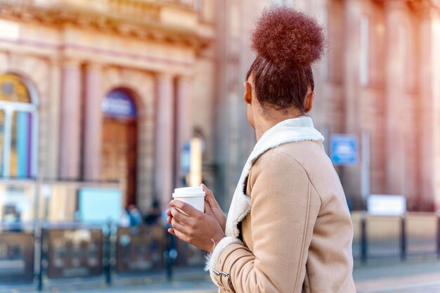 Donna africana detiene una tazza di caffè Una signora bruna riccia sorridente in maglione marrone in attesa di un tram