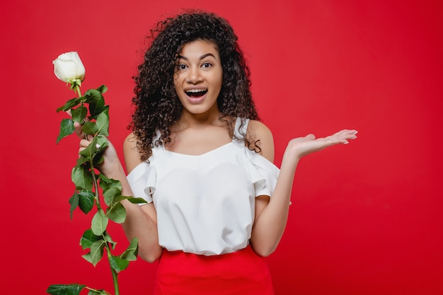 Donna africana emozionante sorpresa con la rosa di bianco isolata sopra rosso