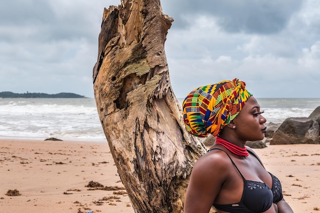 Donna africana ghanese con un copricapo colorato seduta su un tronco di palma rotto
