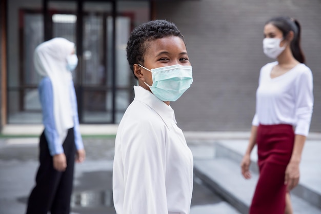 Donna africana nera sorridente felice che indossa una maschera facciale osservando il nuovo stile di vita normale di distanza sociale mantenendo la distanza dalle altre persone in pubblico