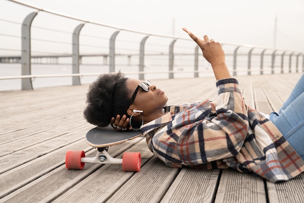 Donna africana premurosa con lo smartphone serio che si trova sullo skateboard sullo spazio urbano della banchina della città scontroso