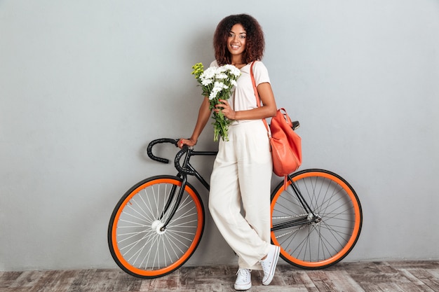 Donna africana sorridente che posa con la bicicletta, lo zaino ed i fiori
