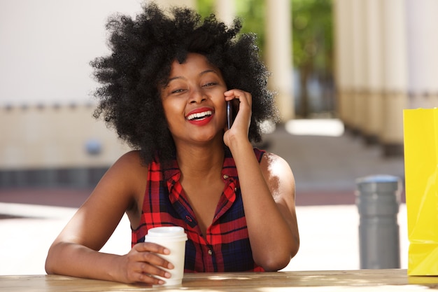 Donna africana sorridente sul cellulare con caffè fuori