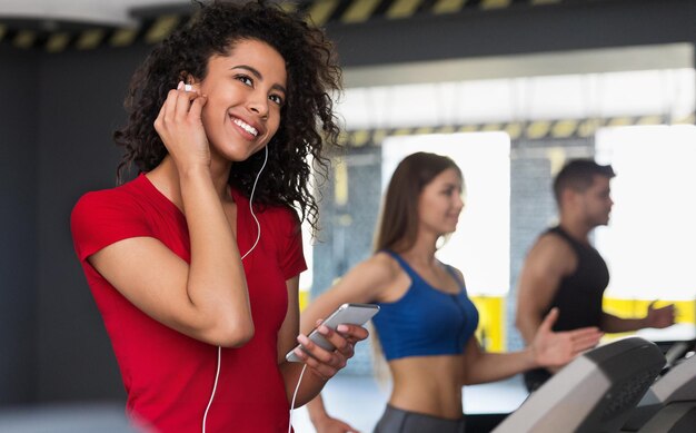 Donna afro-americana in cuffia che corre sul tapis roulant in palestra, sogna e sorride