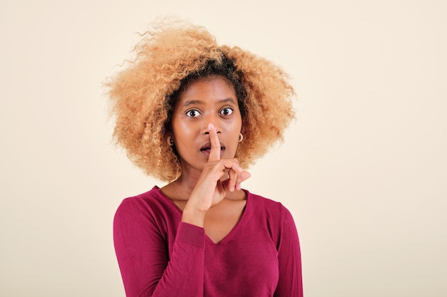 Donna afro che sembra sorpresa mentre mette un dito sulla bocca facendo un silenzio o un gesto segreto su uno sfondo isolato.