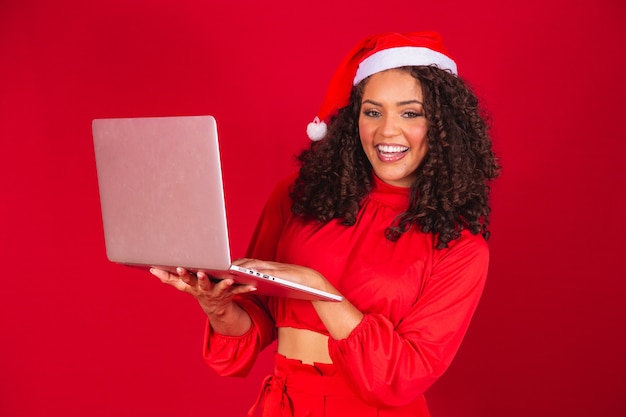 Donna afro con berretto di babbo natale utilizzando laptop su sfondo rosso.