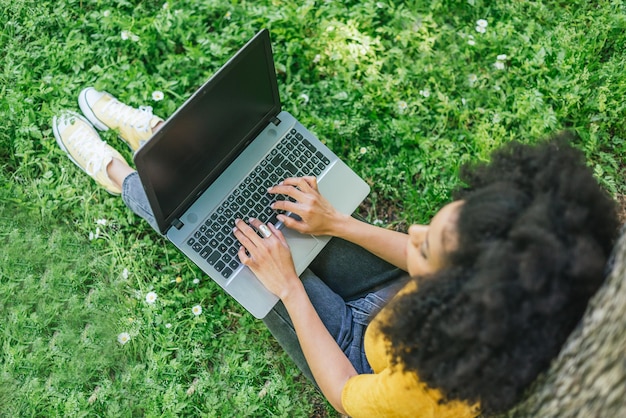 Donna afro digitando su un computer portatile in un giardino. Particolare delle mani sulla tastiera. Messa a fuoco selettiva.