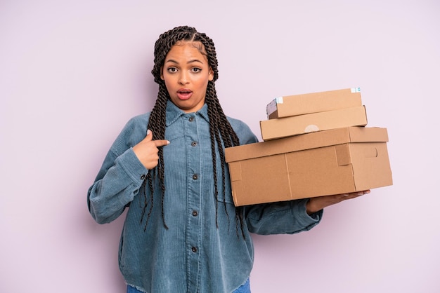 Donna afro nera che sembra scioccata e sorpresa con la bocca spalancata, che indica se stessa. concetto di scatola di pacchetti