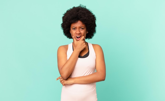 donna afro nera con la bocca e gli occhi spalancati e la mano sul mento, sentendosi spiacevolmente scioccata, dicendo cosa o wow